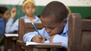 Child in a classroom writing in a notebooks with a pencil in hand. 