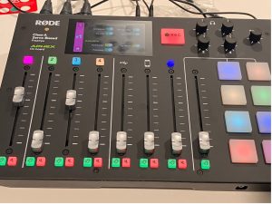 The Rodecaster system is a black base unit approximately 40cm wide with 8 audio sliders, 8 light up effect buttons on the right and a touch screen menu. Wirese protrude from the rear.